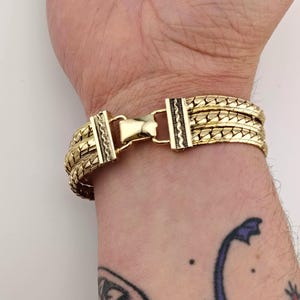 May include: Gold bracelet with a woven design. The bracelet has a secure clasp with decorative detailing. The bracelet is worn on a wrist, showing the intricate pattern and the warm colour of the metal.