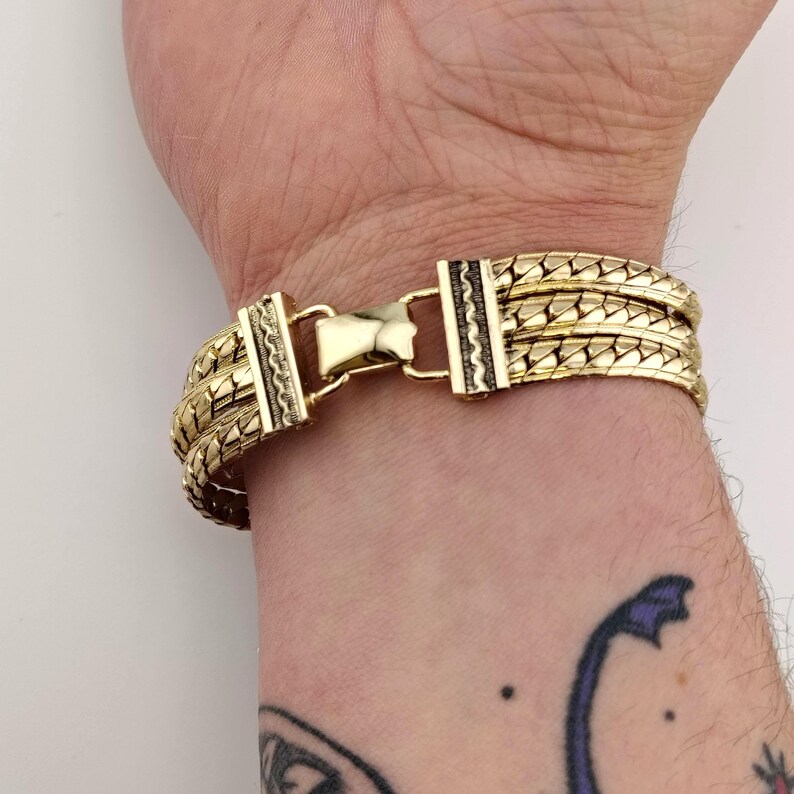 May include: Gold bracelet with a woven design. The bracelet has a secure clasp with decorative detailing. The bracelet is worn on a wrist, showing the intricate pattern and the warm colour of the metal.