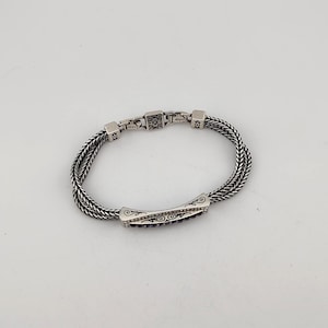 K&ouml;nnte beinhalten: Ein silbernes Armband mit geflochtenem Kettenmuster. Das Armband hat ein rechteckiges Mittelteil mit dunkelvioletten Steinen und dekorativen Wirbeln. Der Verschluss hat ein quadratisches Design mit kleinen schwarzen Steinen. Es ist ein Schmuckst&uuml;ck.