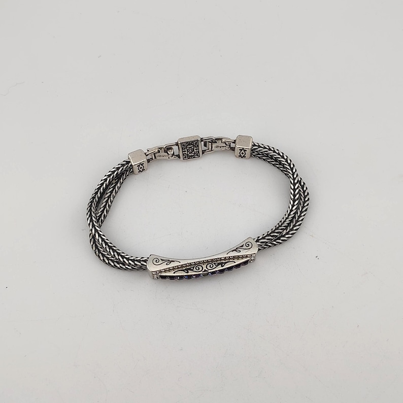 K&ouml;nnte beinhalten: Ein silbernes Armband mit geflochtenem Kettenmuster. Das Armband hat ein rechteckiges Mittelteil mit dunkelvioletten Steinen und dekorativen Wirbeln. Der Verschluss hat ein quadratisches Design mit kleinen schwarzen Steinen. Es ist ein Schmuckst&uuml;ck.