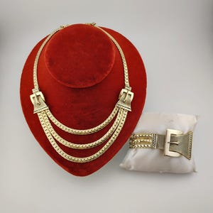May include: A gold-coloured necklace with three cascading strands and a matching bracelet. The necklace features buckle-shaped clasps. The bracelet is on a small white cushion. The jewellery is displayed on a red velvet stand.