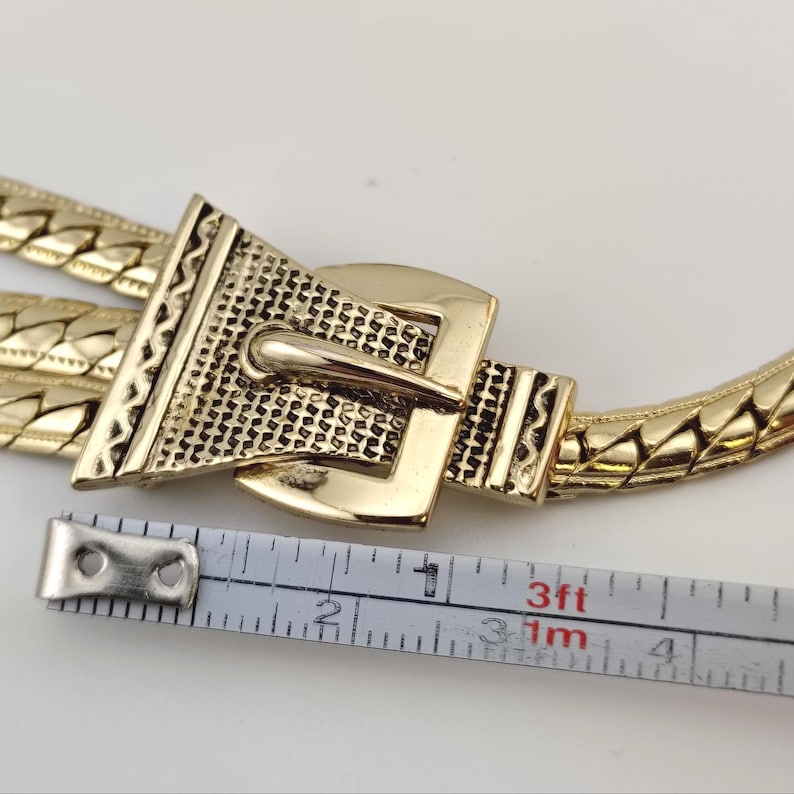 May include: Gold-coloured belt featuring a textured buckle and woven chain. The buckle has a geometric design with a pin closure. A measuring tape shows the belt's length.
