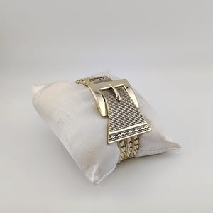 May include: Gold-coloured bracelet with a large buckle design. The bracelet has a woven chain band and a rectangular buckle with a textured pattern. The jewellery is displayed on a white satin pillow against a white background.