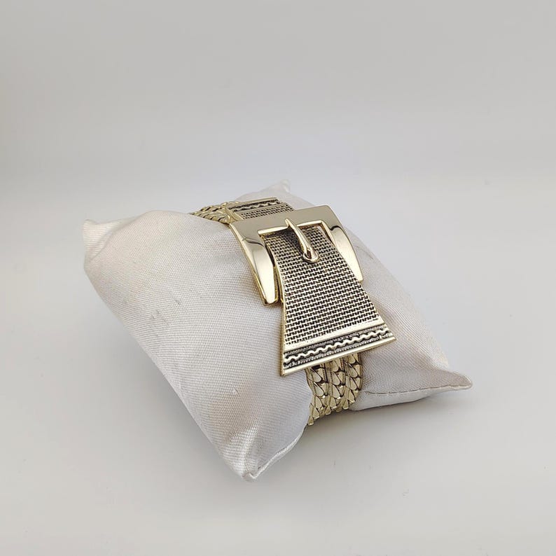 May include: Gold-coloured bracelet with a large buckle design. The bracelet has a woven chain band and a rectangular buckle with a textured pattern. The jewellery is displayed on a white satin pillow against a white background.