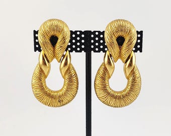 May include: A pair of gold-coloured earrings with a textured, rope-like design. Each earring has a looped top and a teardrop-shaped bottom, connected by a smaller, curved element. The earrings are displayed on a black earring stand against a white background.