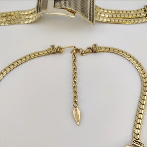 May include: Gold-coloured necklace and bracelet set. The necklace has a chain design with an adjustable chain and a leaf-shaped tag. The bracelet has a geometric design with a textured pattern. Both pieces are on a white background.