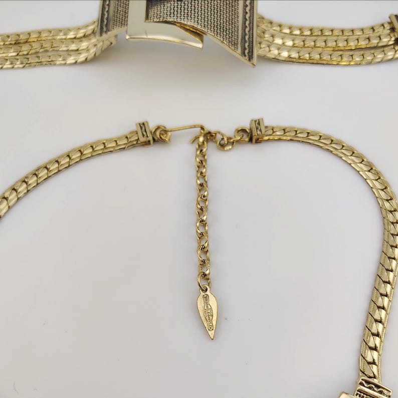 May include: Gold-coloured necklace and bracelet set. The necklace has a chain design with an adjustable chain and a leaf-shaped tag. The bracelet has a geometric design with a textured pattern. Both pieces are on a white background.
