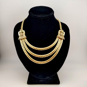 May include: Gold-coloured necklace with a three-strand design. The necklace features decorative buckle-shaped clasps on each end. The necklace is displayed on a black mannequin bust.