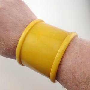 May include: A wide, solid yellow cuff bracelet with a smooth, glossy finish. The bracelet features a curved, cylindrical design that wraps around the wrist, characterised by raised, rounded edges on both the top and bottom of the band.