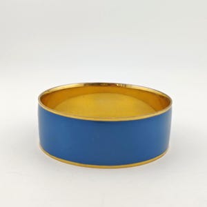 以下が含まれることがあります： 鮮やかなブルーのエナメルバンドが付いた、金色の金属製バングルブレスレット。ブレスレットの内側は金色です。ブレスレットはシンプルでエレガントなデザインです。