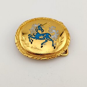 May include: An oval gold-tone belt buckle featuring a central inlay of a blue and white unicorn. The surface of the buckle is decorated with an engraved leaf pattern around the border and a rope-style edge detail surrounding the unicorn design.