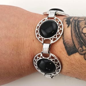 May include: Silver-toned bracelet featuring three oval, black gemstone settings. Each setting is framed by an openwork, geometric design. The bracelet is connected by decorative links and is worn on a wrist.