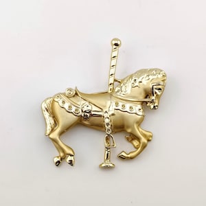 May include: A gold-colored carousel horse brooch. The horse is in a trotting pose, with detailed saddle and bridle. The brooch has a post and clasp closure. The design is a classic, vintage style.