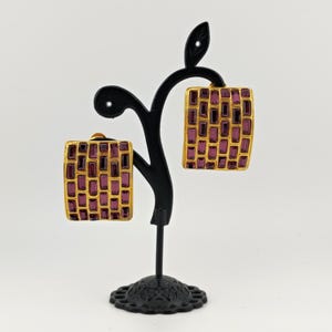 May include: Gold-toned square earrings with a rectangular, amethyst-colored stone pattern. The earrings are displayed on a black jewelry stand against a white background. The earrings have a clip-on style.