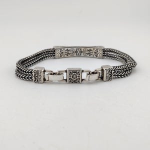 K&ouml;nnte beinhalten: Silberarmband mit geflochtenem Design. Das Armband hat dekorative rechteckige und quadratische Glieder mit komplizierten Mustern. Das zentrale Glied hat ein quadratisches Design und das Armband hat einen sicheren Verschluss. Es ist ein Schmuckst&uuml;ck.