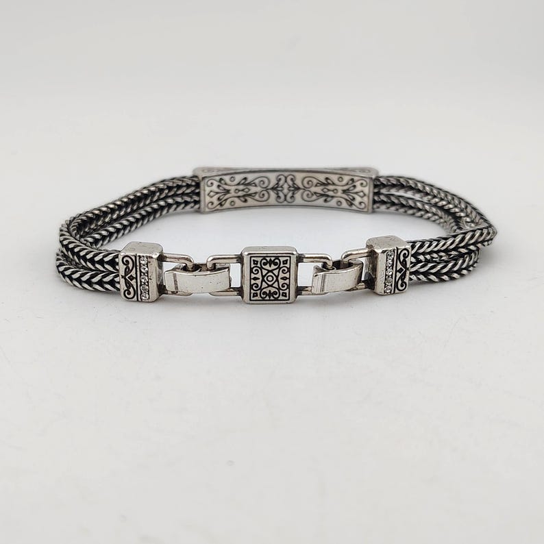 K&ouml;nnte beinhalten: Silberarmband mit geflochtenem Design. Das Armband hat dekorative rechteckige und quadratische Glieder mit komplizierten Mustern. Das zentrale Glied hat ein quadratisches Design und das Armband hat einen sicheren Verschluss. Es ist ein Schmuckst&uuml;ck.