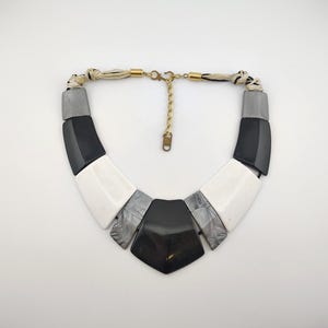 May include: Geometric statement necklace featuring black, white, and gray rectangular and trapezoid-shaped elements. The necklace has a gold-tone chain and a woven fabric cord. The design is bold and modern, suitable for adding a pop of color to any outfit.