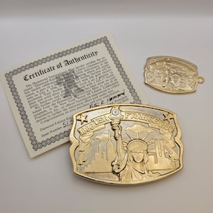 May include: A gold and silver belt buckle with a diamond in the torch of the Statue of Liberty. The buckle is a commemorative piece celebrating the 100th anniversary of the Statue of Liberty. The buckle is inscribed with "1886 USA FRANCE 1986".