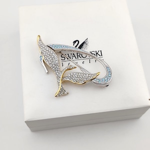 May include: A silver and gold-toned metal brooch shaped like a stylized bird, encrusted with clear crystals and accented with light blue stones. The brooch is positioned on a white box featuring the black Swarovski Jewelry logo and swan icon.