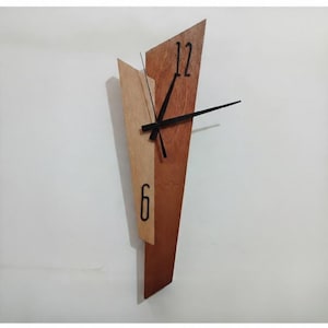 Peut inclure: Une horloge murale dotée d'un cadre en bois asymétrique et allongé, composé de deux nuances de brun distinctes. Le cadran affiche les chiffres noirs 12 et 6, avec des aiguilles noires fines positionnées au centre de la structure en bois.