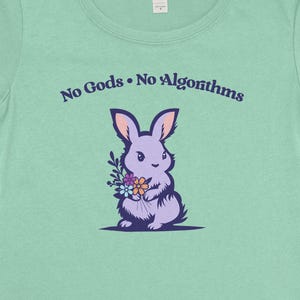 May include: A light green t-shirt featuring a purple bunny holding flowers. The text "No Gods No Algorithms" is arched above the bunny. The shirt has a crew neck and short sleeves.