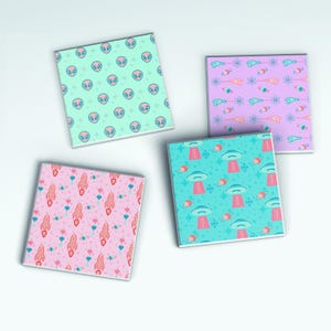 May include: Four square ceramic coasters with colourful patterns. Designs include alien heads, ray guns, rockets, and UFOs. The coasters are in shades of pink, blue, and purple, with white backgrounds. These coasters are perfect for adding a touch of fun to any space.