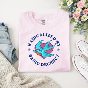 以下が含まれることがあります： 「RADICALIZED BY BASIC DECENCY」の文字が円状に配置され、青い鳥のグラフィックとハートが描かれた、薄いピンク色のスウェットシャツ。白い表面に折りたたまれ、白いスニーカーと花のブーケが添えられています。