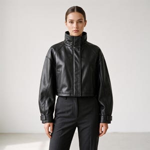 May include: A black cropped jacket made of a smooth material with a high stand-up collar, long sleeves with adjustable cuffs, and a front placket covering the zipper. The jacket is paired with dark gray trousers featuring a center seam.