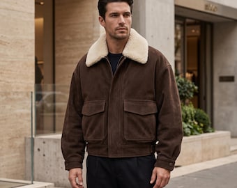 Può includere: Un giubbotto bomber in camoscio marrone scuro con un colletto spesso in montone color crema, chiusura frontale con zip e due ampie tasche con patta sul petto. La giacca presenta maniche lunghe con polsini abbottonati e orlo dritto.