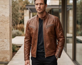 Può includere: Una giacca in pelle marrone con colletto alla coreana e chiusura con bottone a pressione, cerniera frontale, due tasche sul petto con zip e polsini con zip. La giacca è indossata sopra un maglione grigio scuro e pantaloni neri.