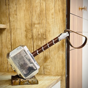 Op de afbeelding: Een zilverkleurige hamerreplica met een bruin, met leer omwikkeld handvat en een bruine leren polsband. De hamerkop is voorzien van ingewikkelde gegraveerde patronen en een handtekening met de tekst THE KID in het metalen oppervlak.