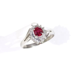 May include: A silver-toned ring featuring a central oval-cut deep red gemstone surrounded by a halo of small, clear, round-cut stones. The band is smooth and tapers toward the setting, which has a prong-style mount for the center stone.