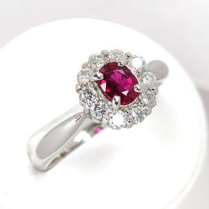 May include: A silver-toned ring featuring a central oval-cut deep red ruby gemstone. The ruby is surrounded by a halo of twelve small, round-cut clear white diamonds set in a circular arrangement. The band is smooth, polished, and metallic.