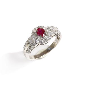 May include: A silver-toned ring featuring a central oval-cut red ruby gemstone surrounded by a halo of small round white diamonds. The band is split into multiple rows, each encrusted with additional small white diamonds along the sides.
