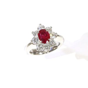 May include: A silver-toned ring featuring a central oval-cut deep red ruby gemstone. The ruby is surrounded by a cluster of eight round-cut clear white diamonds arranged in a floral pattern, set on a polished metallic band with a smooth finish.