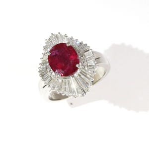 May include: A silver-toned ring featuring a large, oval-cut deep red gemstone at the center. The central stone is surrounded by a halo of numerous clear, baguette-cut stones arranged in a fan-like pattern, set on a polished metal band.