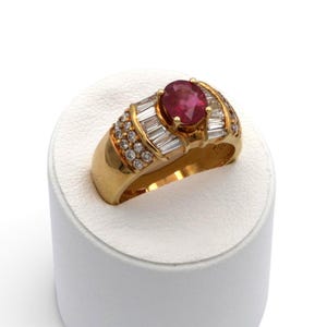 May include: A gold ring featuring a central oval-cut red gemstone held by four prongs. The band is accented with baguette-cut diamonds flanking the center stone and rows of small round-cut diamonds along the sides of the wide, polished gold band.