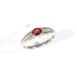 May include: A silver-toned ring featuring a central oval-cut red gemstone flanked by a row of small, clear, round-cut stones set into the band on both sides. The metal band has a polished finish and a smooth, tapered design.