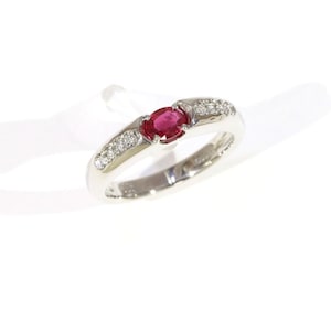 May include: A silver-toned ring featuring a central oval-cut red gemstone held in a prong setting. The band is accented with small, clear, round-cut stones embedded along the shoulders on both sides of the primary red stone.
