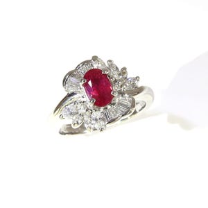 May include: A silver-toned ring featuring a central oval-cut red ruby gemstone. The ruby is surrounded by a cluster of clear, faceted diamonds, including marquise-cut stones and baguette-cut stones arranged in a fan-like pattern on the band.