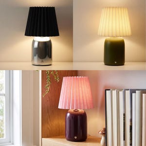 May include: A collection of three table lamps, each featuring a cylindrical base and a pleated lampshade. The lamps are shown in three color variations: a chrome base with a black shade, an olive green base with a white shade, and a dark purple base with a pink shade.