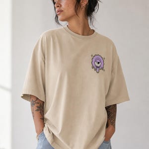 Könnte beinhalten: Ein beiges Oversize-T-Shirt mit einer Grafik auf der linken Brust. Das Motiv zeigt eine violette, schmelzende Kreisform mit einem Auge in der Mitte und kleinen Sternakzenten. Das T-Shirt hat einen Rundhalsausschnitt und kurze Ärmel, getragen zu Jeans.
