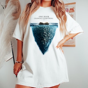 Könnte beinhalten: Ein cremefarbenes T-Shirt mit kurzen Ärmeln und einer Grafik eines aufgeschlagenen Buches auf dunkelblauem Wasser, unter dem sich eine große, untergetauchte Masse gestapelter Bücher befindet. Der Text ONE BOOK CHANGES EVERYTHING steht darüber.