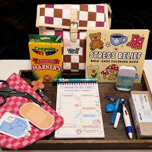 May include: A collection of items on a wooden tray including a brown and white checkered bag, a box of Crayola markers, a Stress Relief coloring book, a Nurse To Do List notepad, black scissors, a pink checkered cloth, pens, and a hand sanitizer.