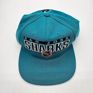 Op de afbeelding: Een groenblauwe honkbalpet met de geborduurde tekst SAN JOSE SHARKS in zwarte en witte letters op de voorkant. Onder de tekst staat een klein oranje en zwart NHL-schildlogo. De pet heeft een gestructureerde kroon en een platte klep.