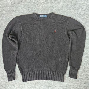 May include: A dark charcoal gray long-sleeve crewneck sweater featuring a ribbed collar, cuffs, and hem. The sweater has a small red embroidered logo on the left chest and a blue Polo by Ralph Lauren label inside the neckline collar.
