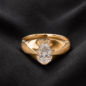 May include: A polished yellow gold ring featuring a single pear-shaped faceted clear gemstone set securely within a smooth, tapered metal band that curves upward to frame the stone in a bezel-like setting with a reflective, high-shine finish.