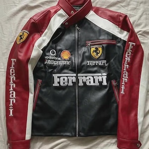 May include: A black, red, and white racing jacket featuring the Ferrari logo, text, and various sponsor emblems including Vodafone, Shell, and Bridgestone. The jacket has a stand-up collar, a full-length front zipper, and zippered pockets.