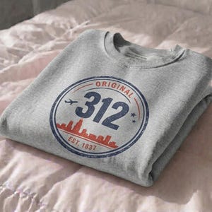 May include: A heather gray crewneck sweatshirt featuring a circular graphic with the text ORIGINAL 312 EST. 1837. The design includes a red city skyline silhouette, a small airplane icon, and two stars, all enclosed within a distressed blue circular border.