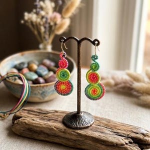 May include: A pair of dangling earrings featuring three stacked, spiraled circular components in vibrant shades of pink, lime green, and orange. The earrings are crafted from coiled thread and hang from gold-toned metal hooks on a dark metal stand.